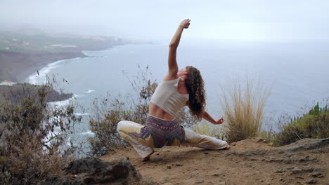 At-the-cliff's-brink,-a-woman-in-warrior-pose-lifts-her-hands,-inhaling-the-sea-breeze-while-embracing-the-yoga-session-amid-ocean-vistas